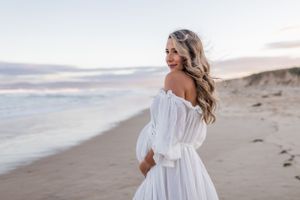 A woman in an off-shoulder white dress poses on a beach during golden hour.