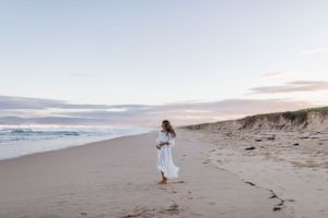 A woman in a white dress walks along a serene beach at sunset.