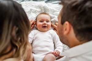 A joyful baby lying between parents on white bedding.