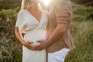 Close up of pregnant belly with hands forming heart shape in sunlit field.