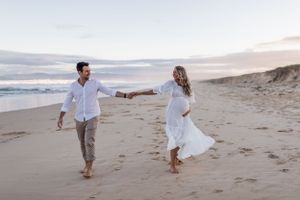 A couple holding hands and dancing on a beach at sunset in white and casual attire.