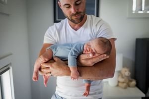 Baby in light blue outfit being cradled in parent's arms in a nursery room.