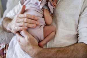 Close-up view of hands gently cradling a sleeping infant swaddled in light fabric.