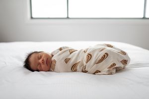 Peaceful sleeping newborn swaddled in rainbow patterned blanket on white bedding.