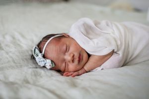 Peaceful sleeping newborn wearing white swaddle and headband on textured bedding.
