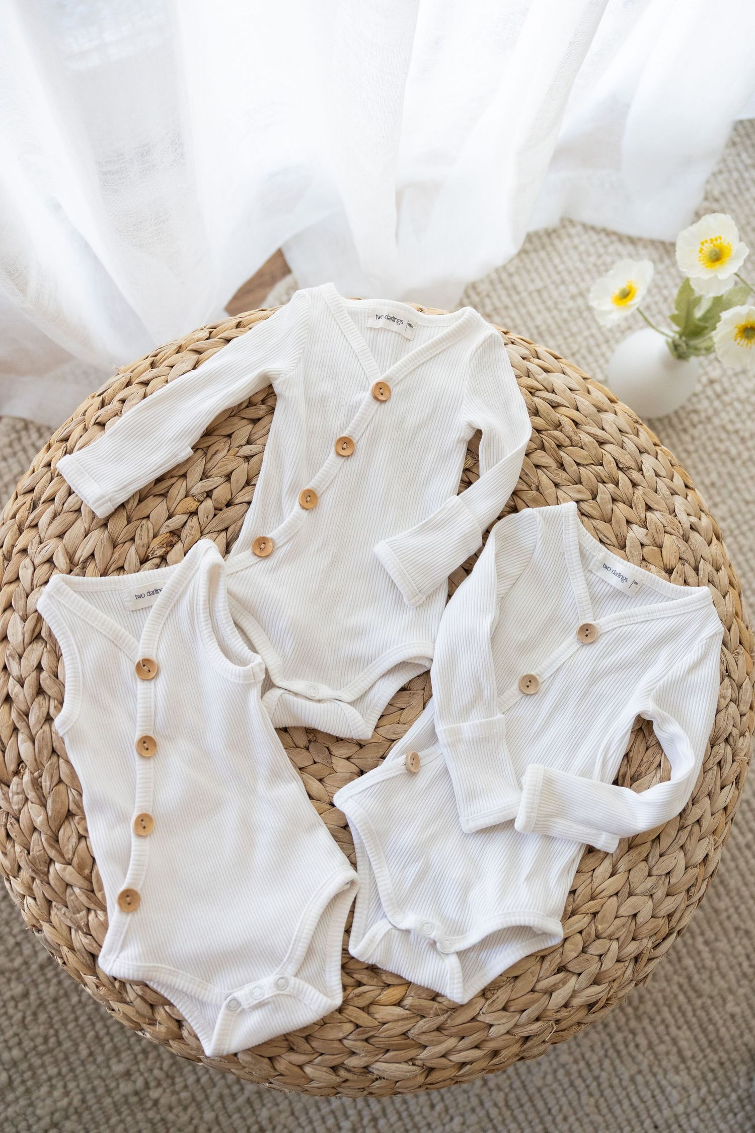 White cotton baby onesies with wooden buttons arranged on a woven rattan mat with delicate white flowers in the background.