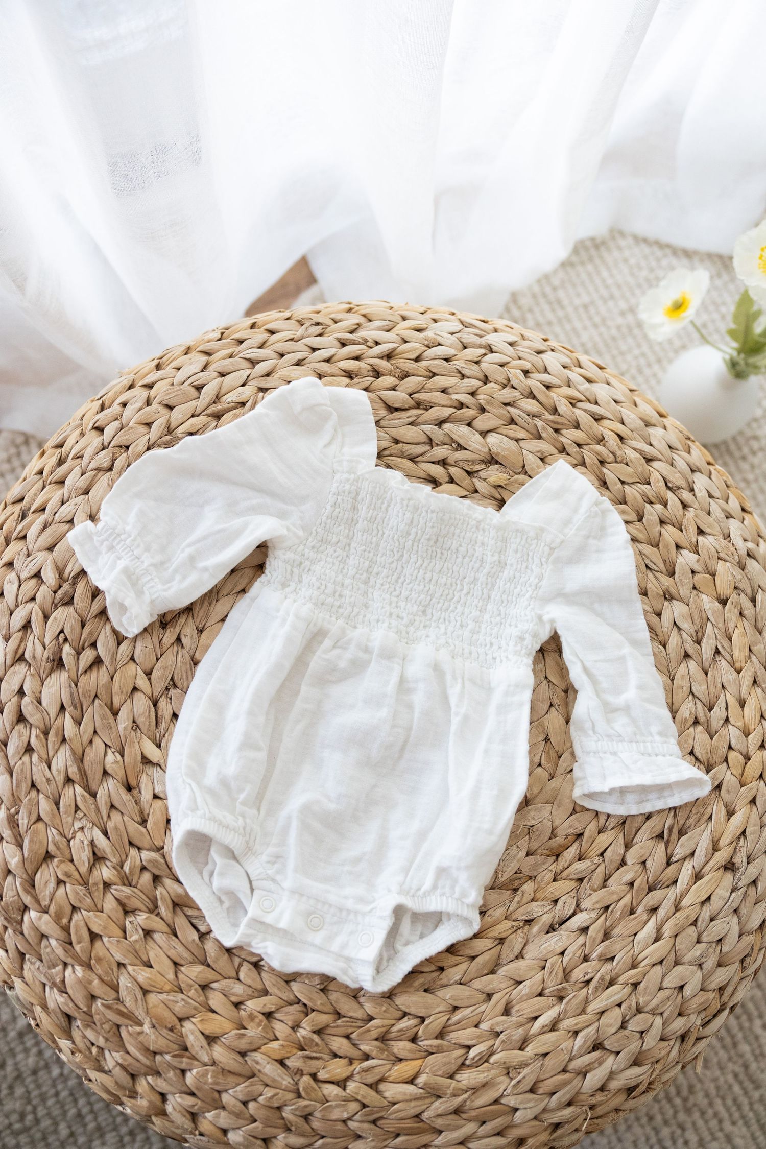 White muslin baby romper with puff sleeves displayed on round woven rattan mat with delicate floral accent.