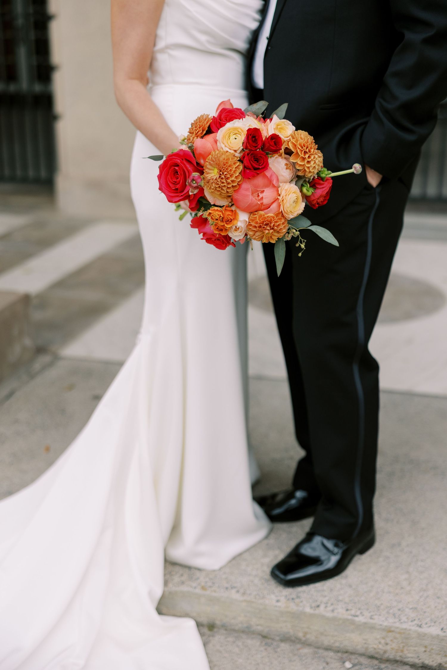 Wedding couple standing on steps with elegant coral and red rose bridal bouquet against white dress and black tuxedo at Hall of Springs.