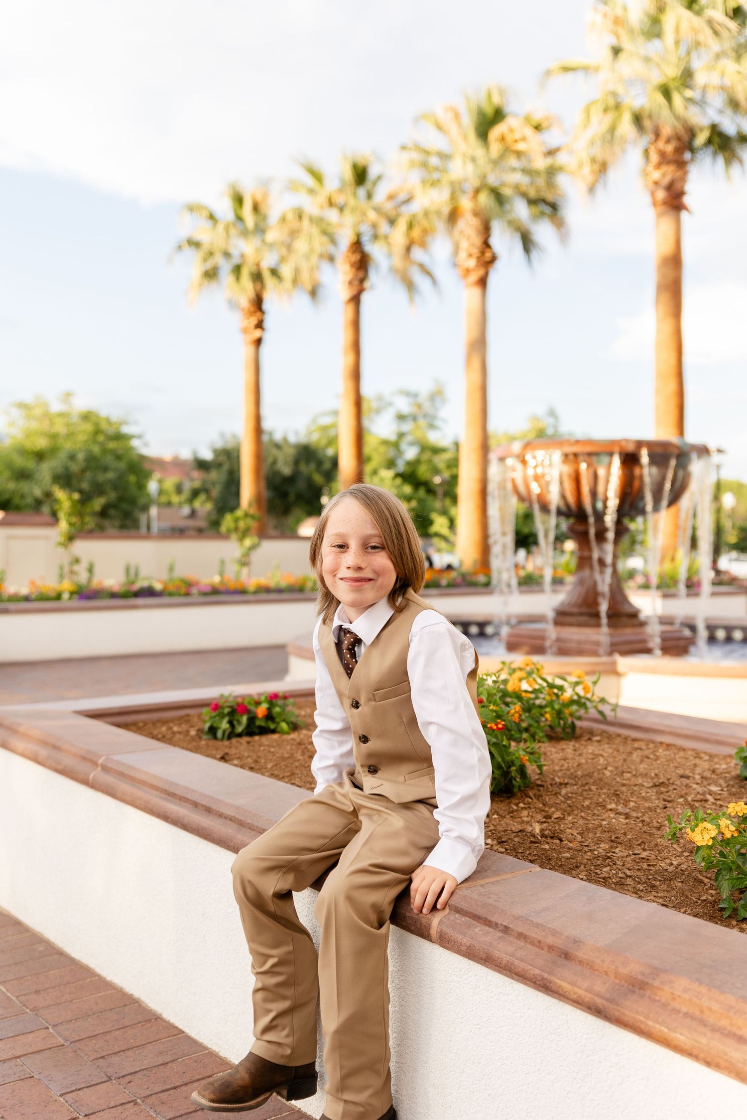 boy sitting in brown suit on the edge of a flower bed with 4 large palm trees lining the background