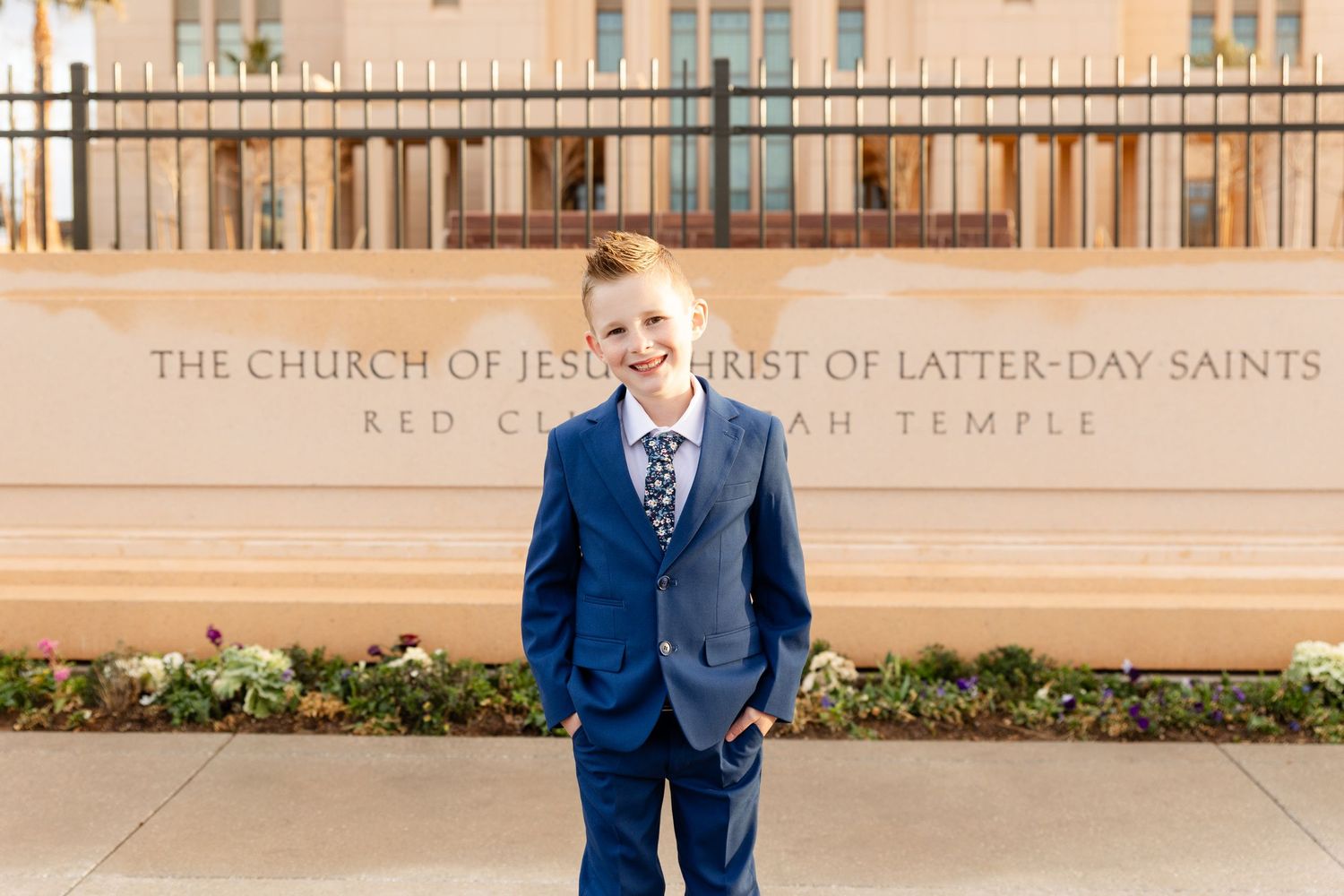 boy in blue suit hands in pockets smiling at the camera with a sign "The Church of Jesus Christ of Latter-day Saints" in the background