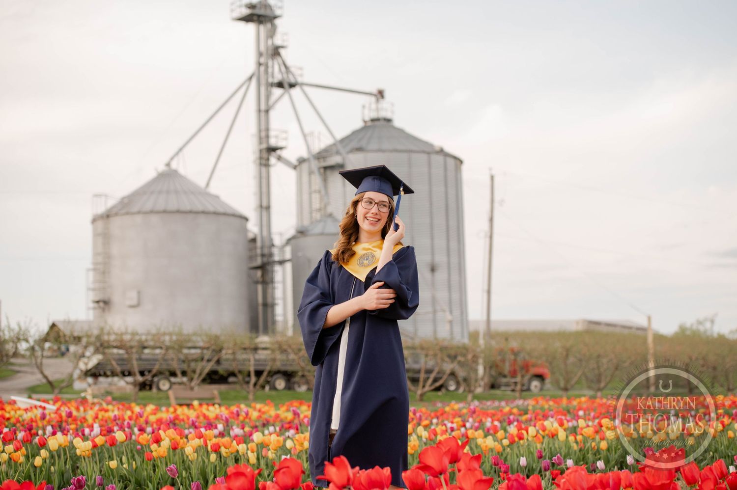 Graduate in navy cap and gown stands in colorful tulip field with grain silos in background.