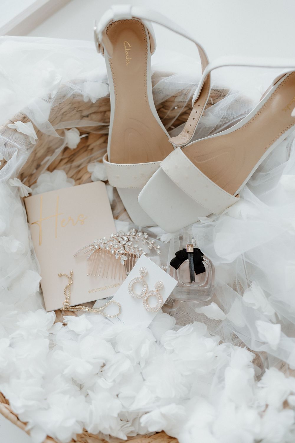 Elegant white bridal accessories including sandals, rings, perfume and invitations arranged on soft white tulle fabric.
