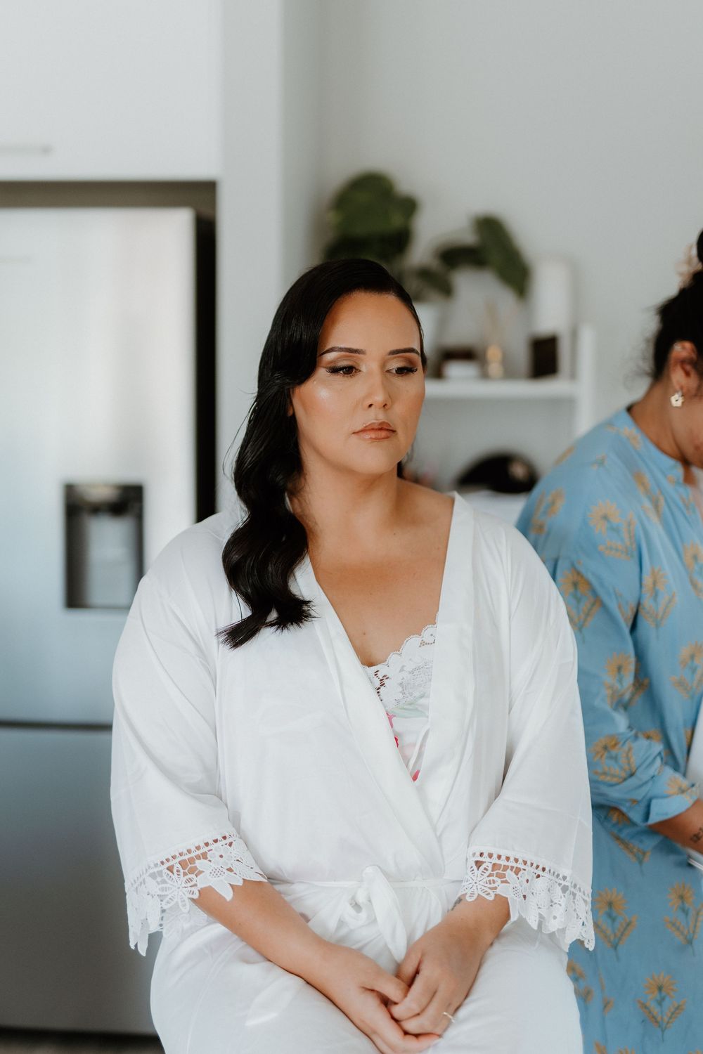 Woman wearing a white lace-trimmed robe sits poised during wedding day preparations in a modern room.