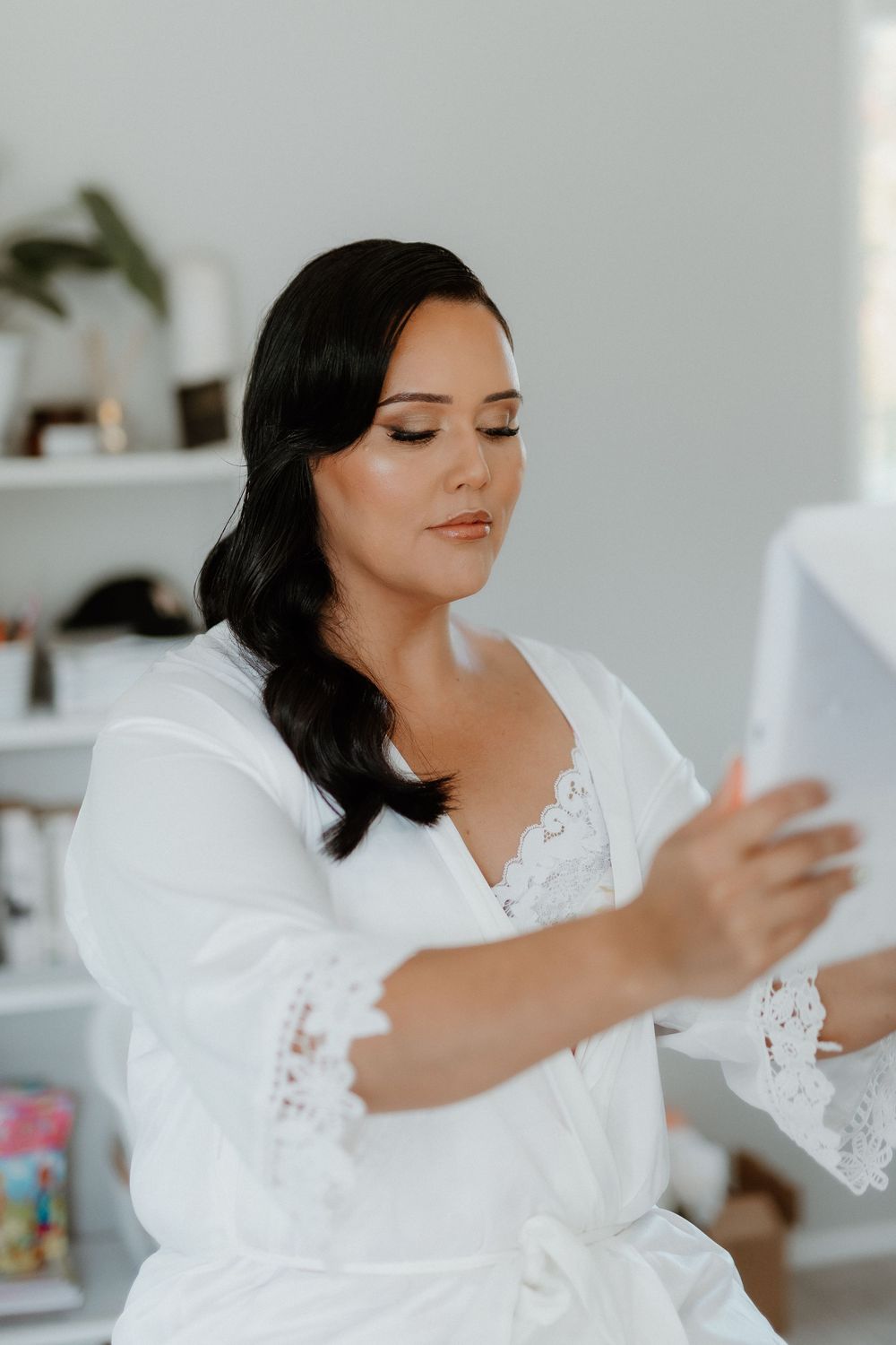 Person wearing white lace robe with long dark hair looks down while holding something in minimalist setting.