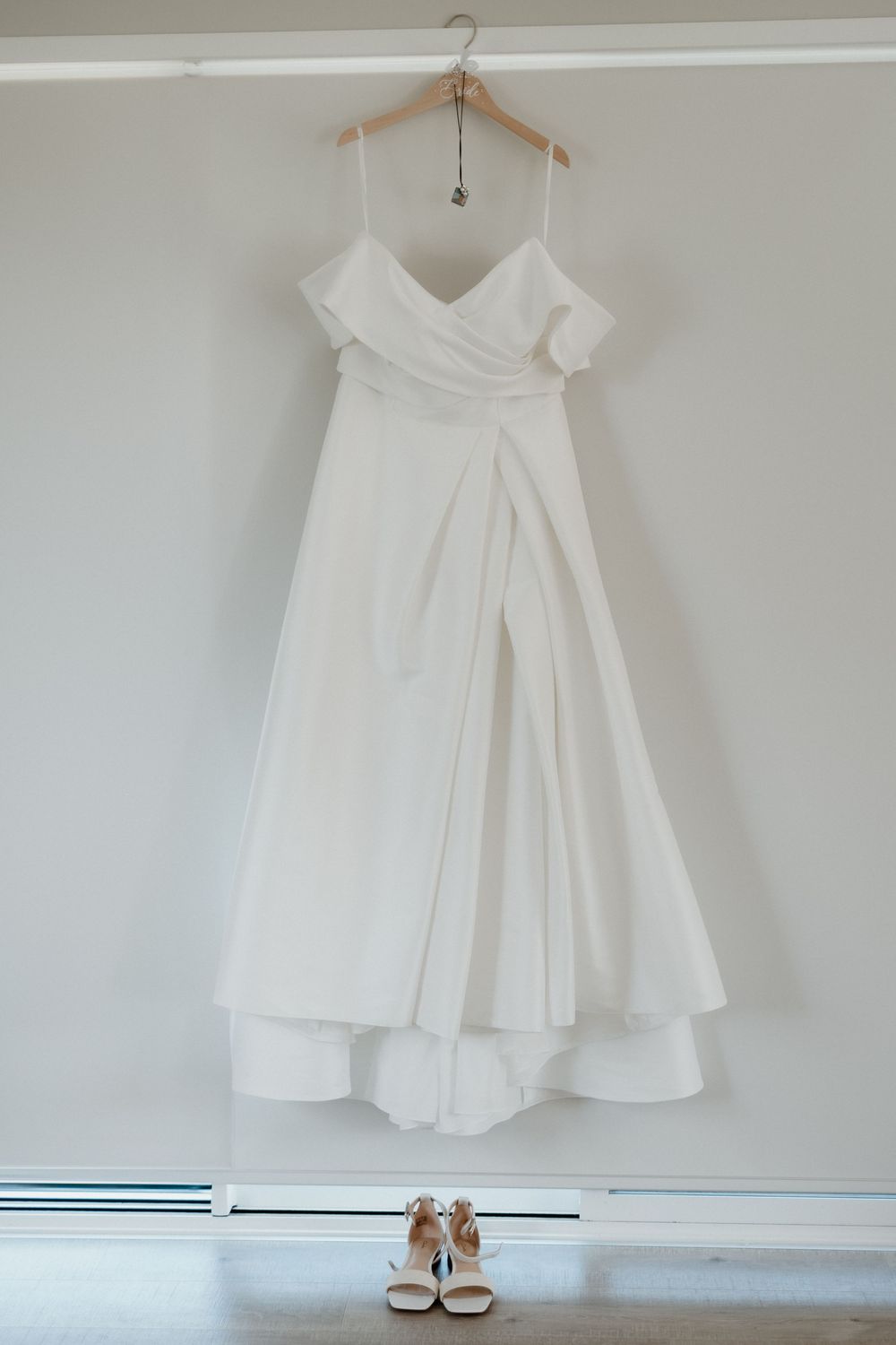 An elegant white wedding dress with off-shoulder design and flowy skirt hangs on wooden hanger above matching bridal shoes.