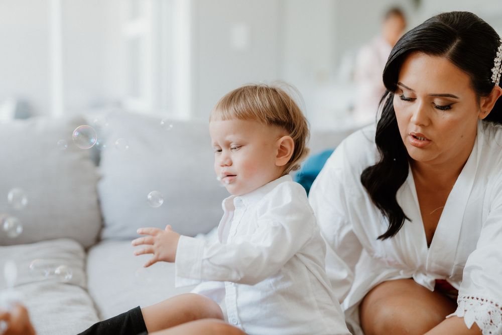 Small child in a white dress excitedly reaches for bubbles while sitting on a couch with an adult in a white robe.