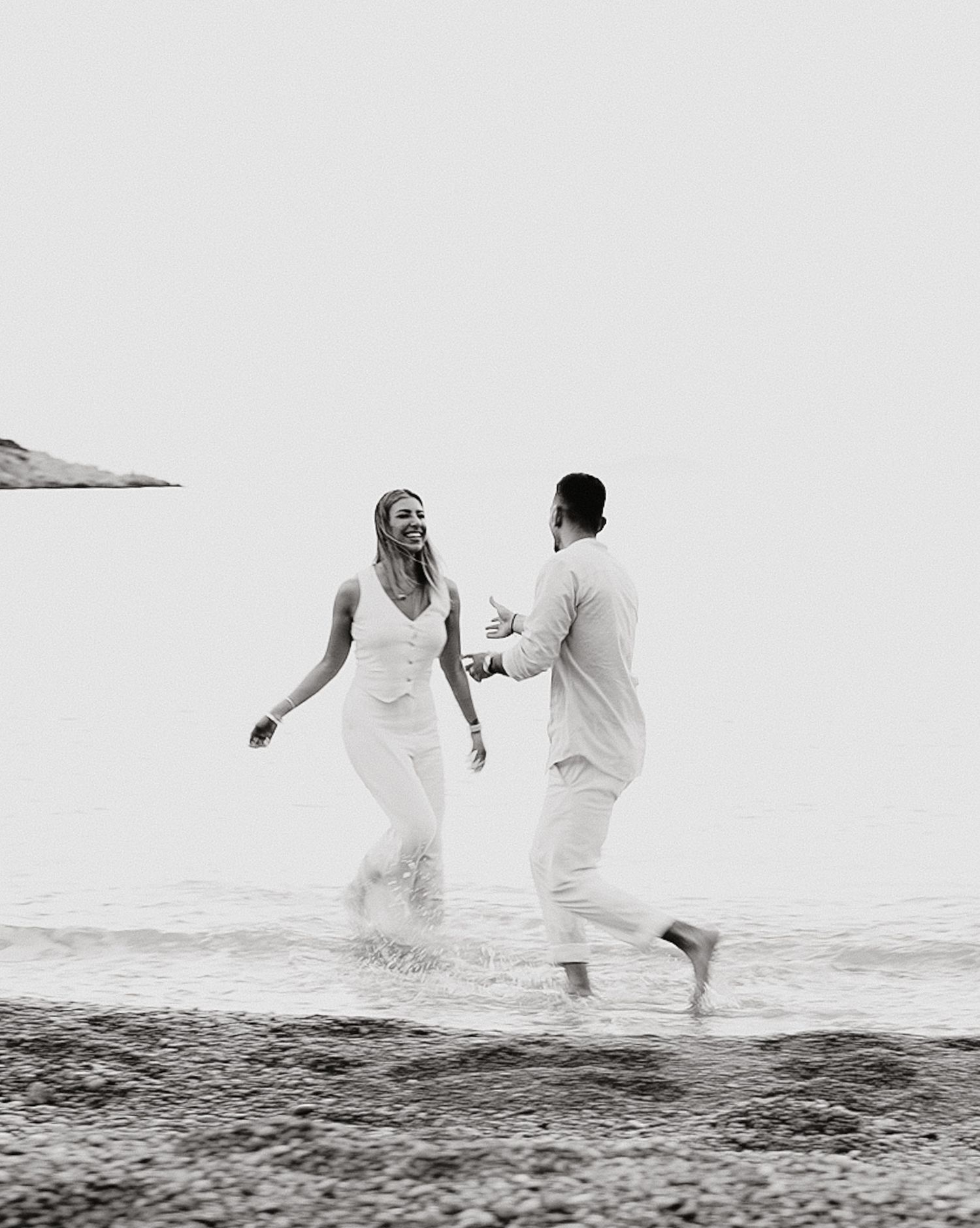 A couple in white clothing runs joyfully through shallow ocean water together in black and white photography.