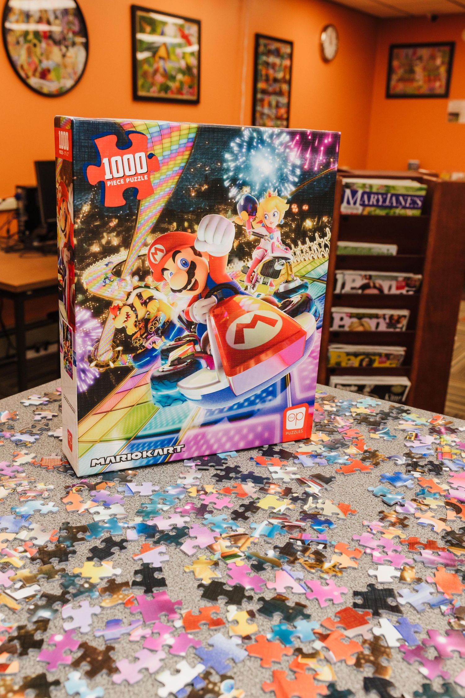A 1000-piece Mario Kart puzzle box sits on a community table covered in scattered colorful puzzle pieces with orange walls in background.