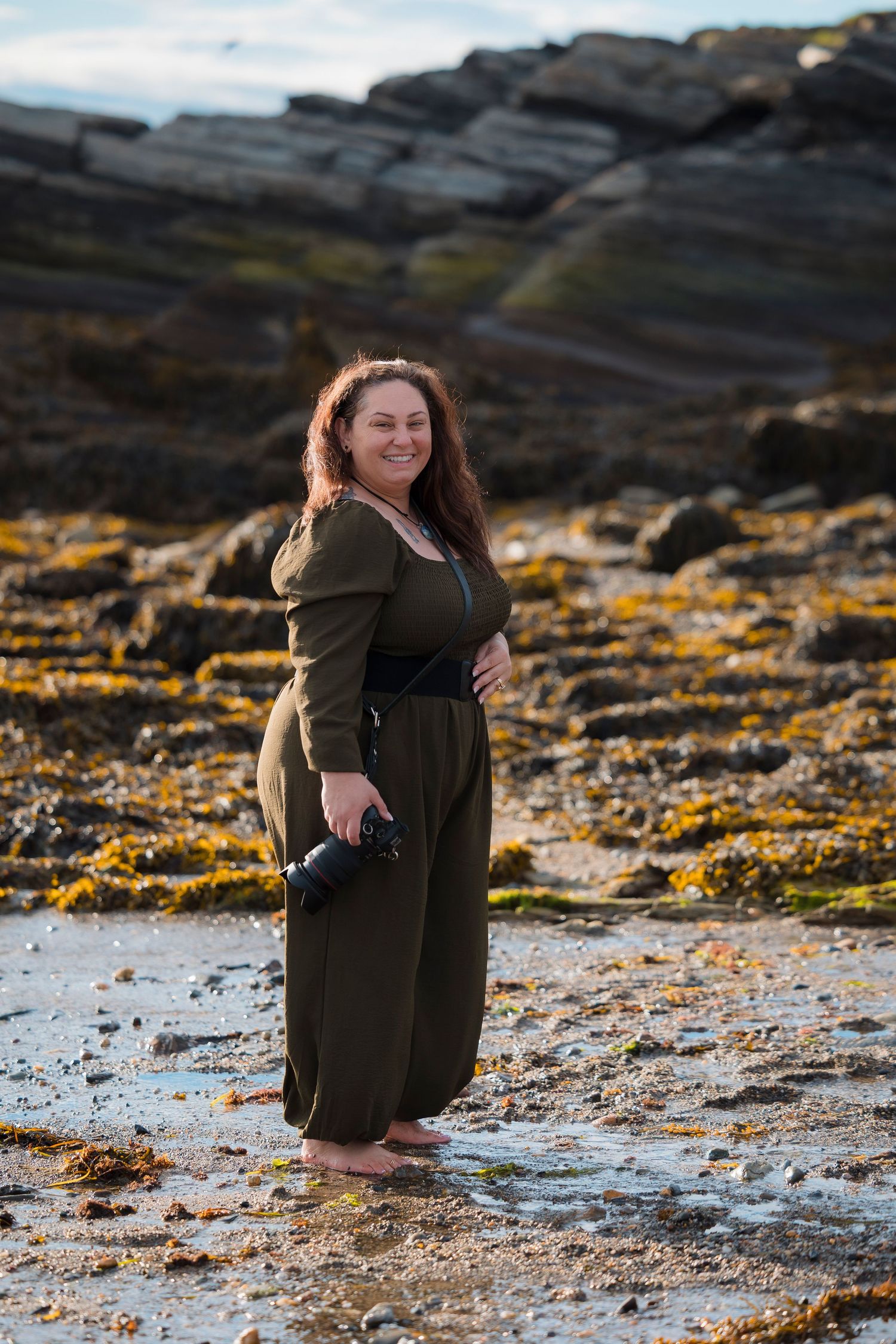 Someone in an olive green dress poses on a rocky coastline with yellow lichen and tidepools at sunset.