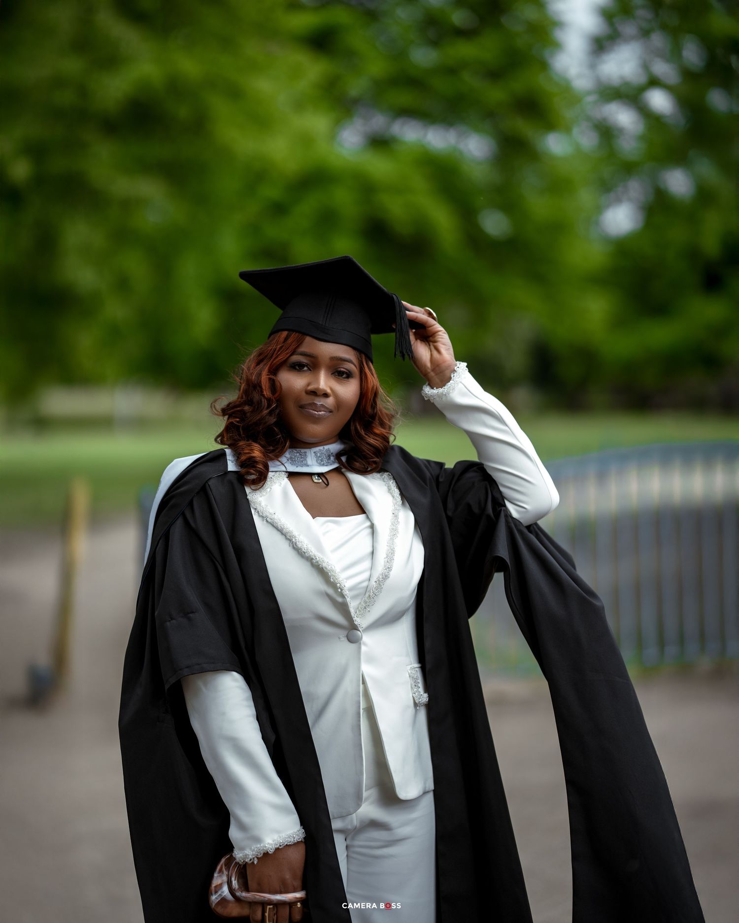 A graduate in white suit and black graduation gown poses outdoors against a blurred green background while adjusting their cap.
