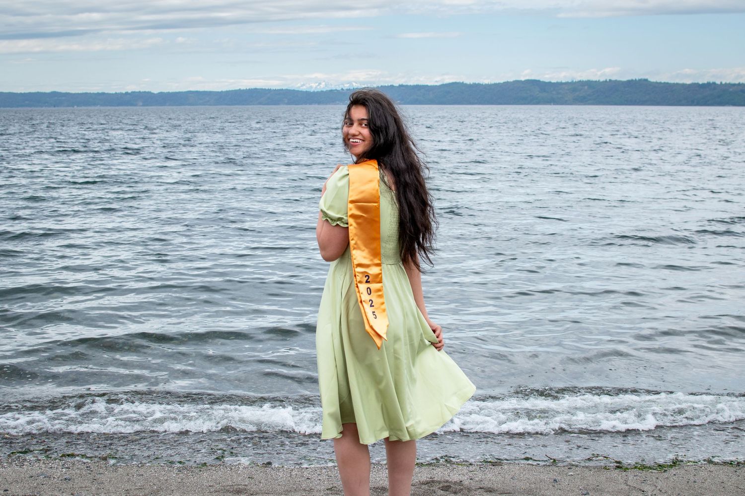 Graduation portrait of senior on the beach in Des Moines, WA, holding a gold 2025 sash. Redondo Park. Senior photography.