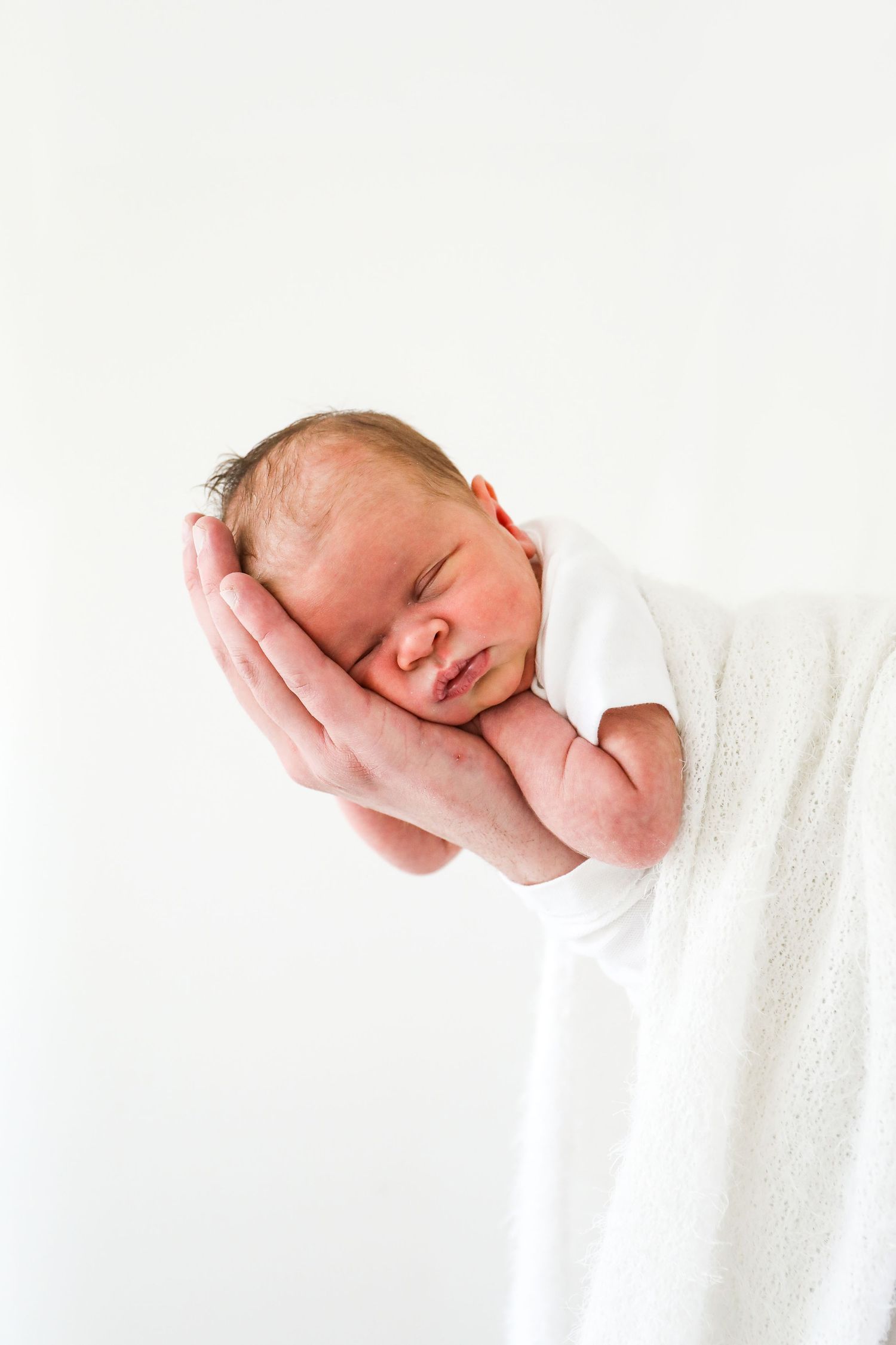 A newborn baby sleeps peacefully while being cradled in dads hands against a white background, during newborn shoot in Nottingham