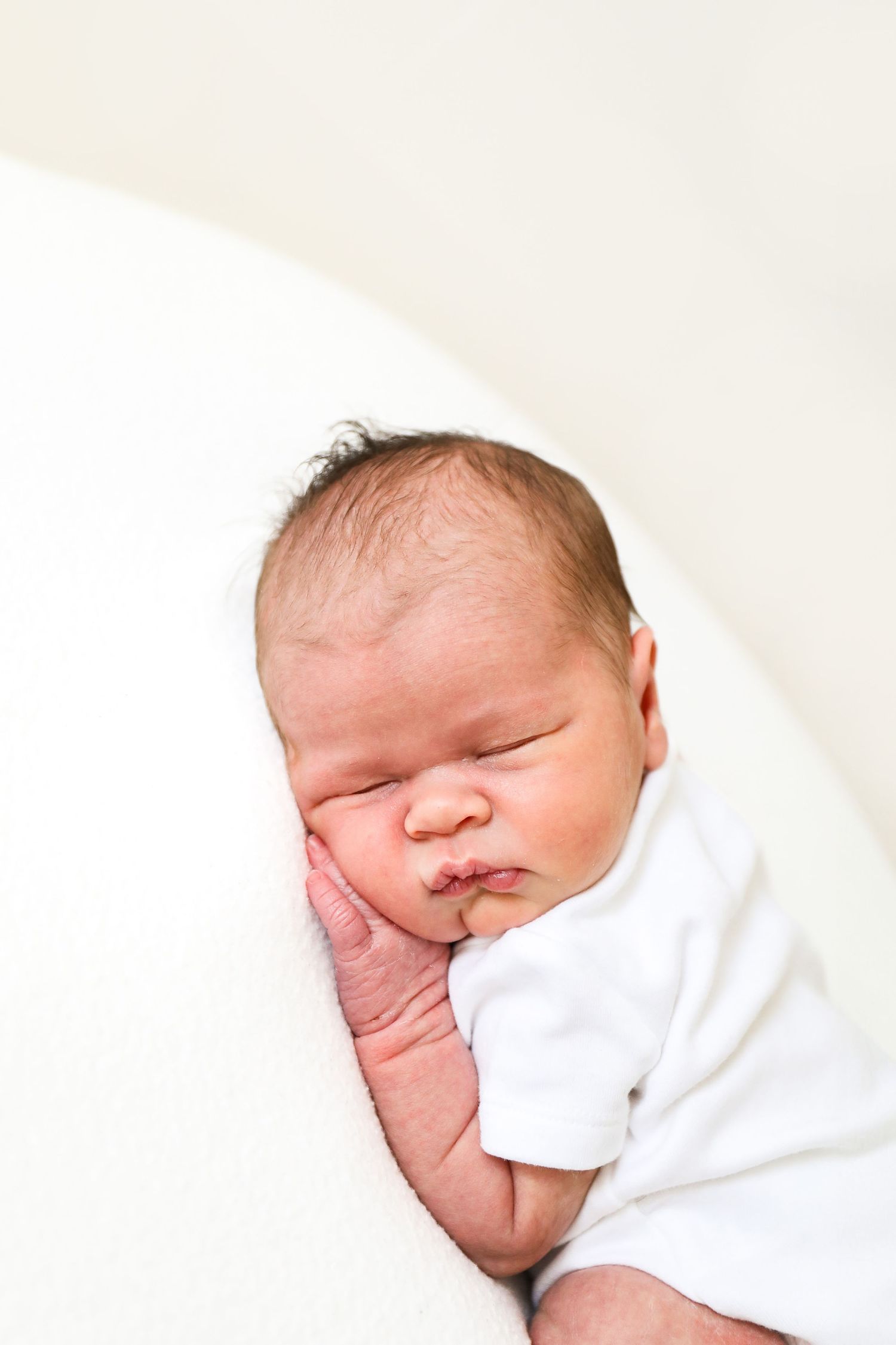 Sleeping newborn baby in white onesie curled up on soft white bedding, during newborn shoot in Nottingham