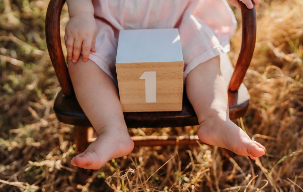 Small bare feet and wooden number one block on a rustic wooden chair during a sunny outdoor photoshoot.