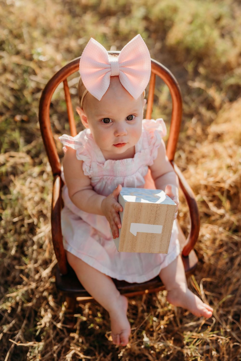 A small figure wearing a pink dress and bow sits in a wooden chair holding a wrapped gift, surrounded by golden grass.