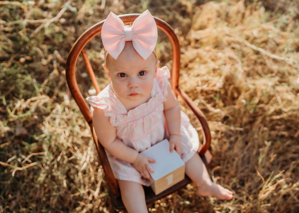 A small child in a pink outfit with a matching bow sits in an antique wooden chair against a natural outdoor background.