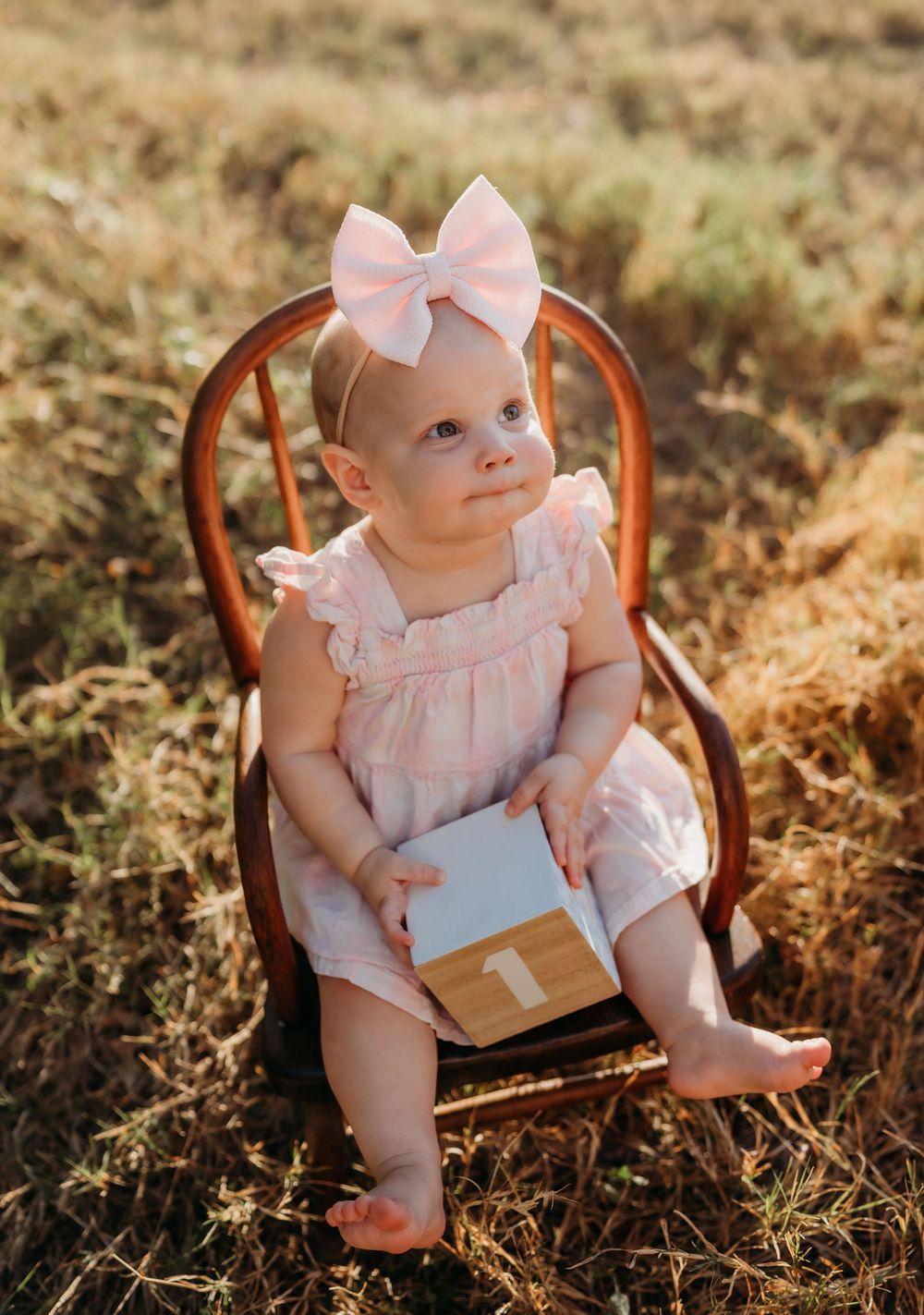 A small baby sits in a wooden chair wearing a pink dress and bow while holding a number block in a golden field.