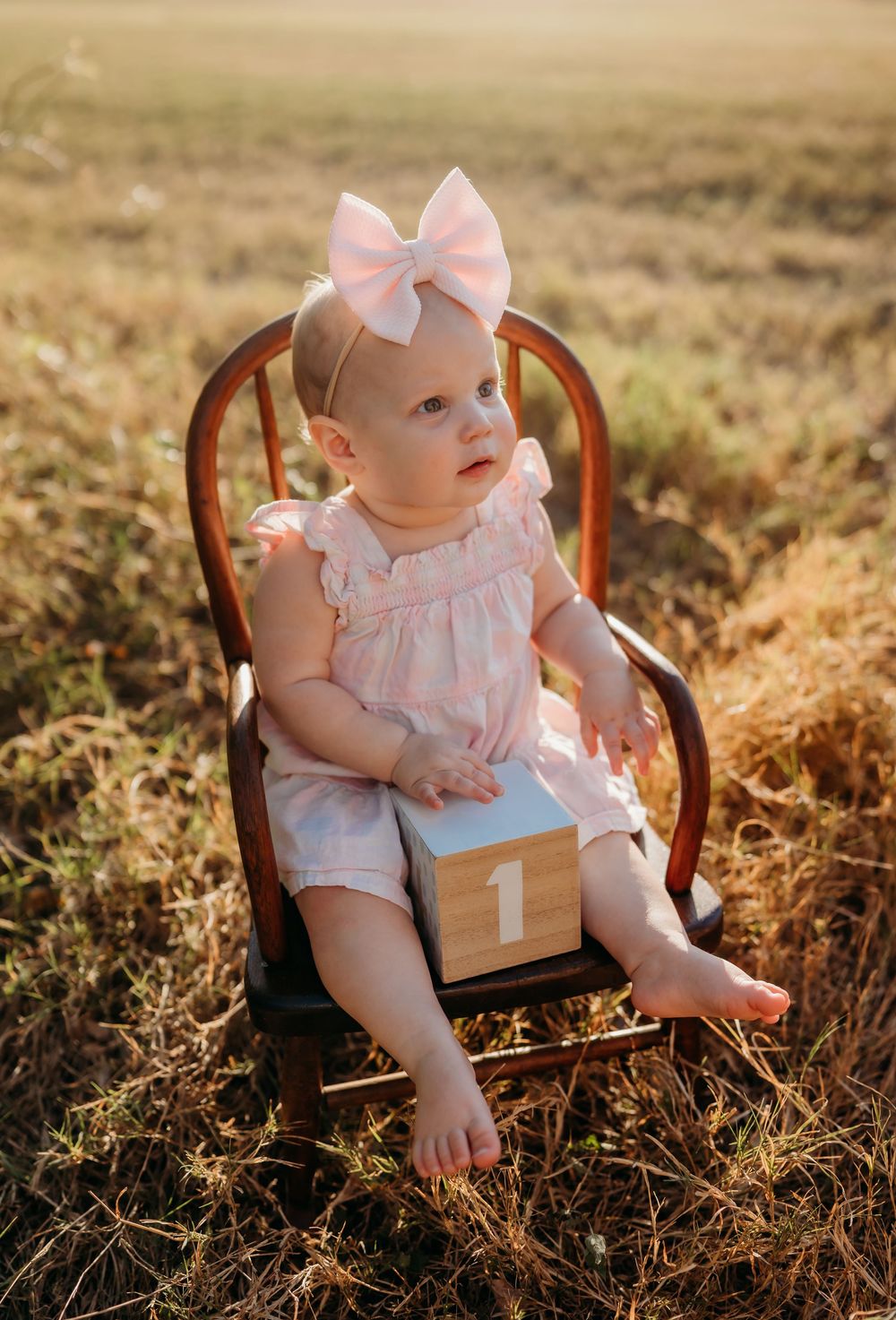 A baby in a pink outfit with bow sits in a wooden chair holding a number 1 block during golden hour.