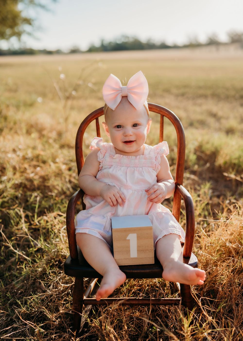 Baby wearing pink bow and dress sits in wooden chair outdoors in golden sunlit field with number one decoration.