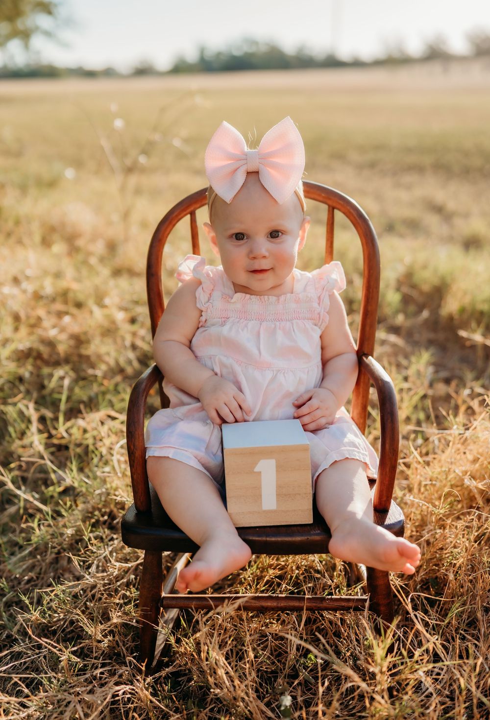 A wooden chair sits in a golden sunlit field with a number one block and pink bow accessories.