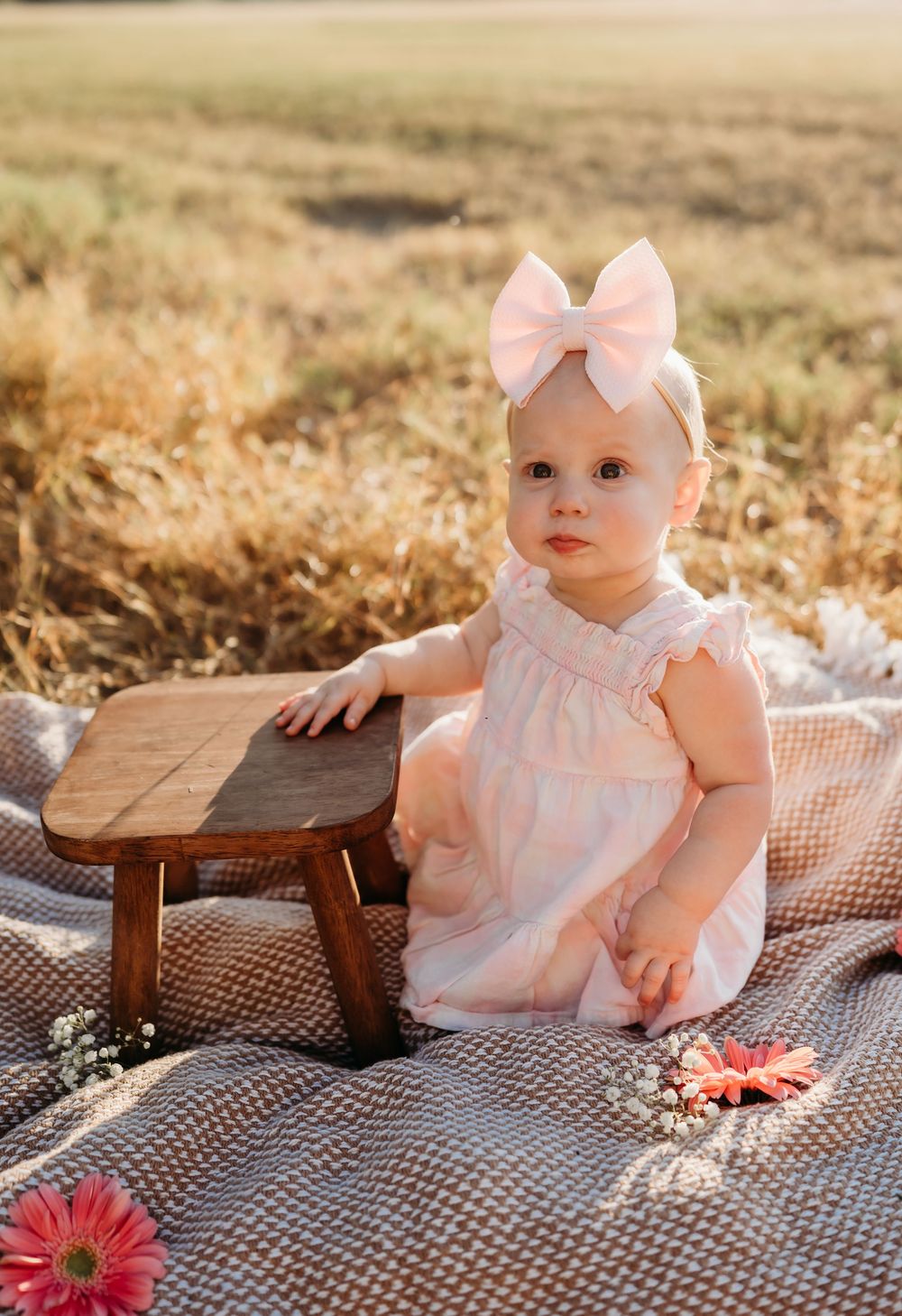 Small child in pink dress and bow sits on picnic blanket with wooden stool in golden field during sunset.
