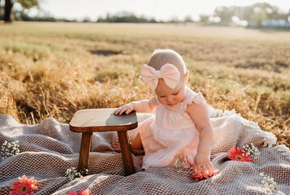 A small figure in a white dress sits on a picnic blanket with a wooden table in a golden wheat field at sunset.
