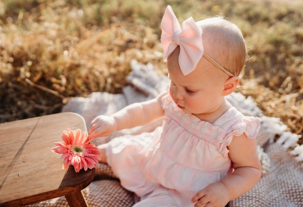 Baby in pink dress with white bow headband sits on blanket looking at coral gerbera daisy flower in sunlit outdoor setting.