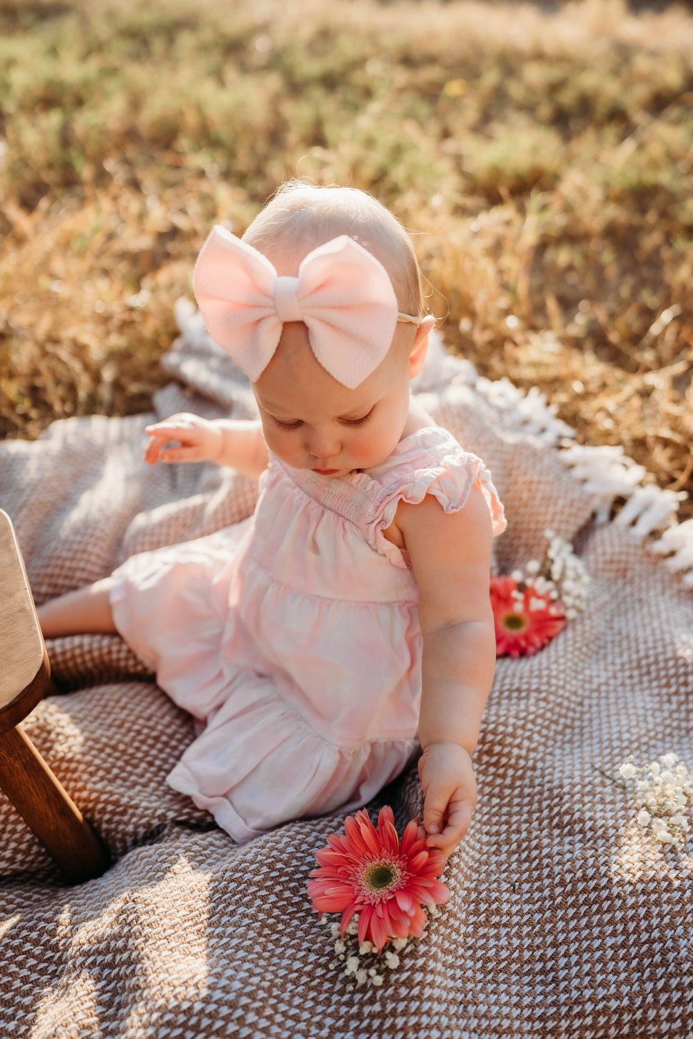 Baby in pink dress and bow sits on gray blanket with red gerbera daisy flower during outdoor photo session.