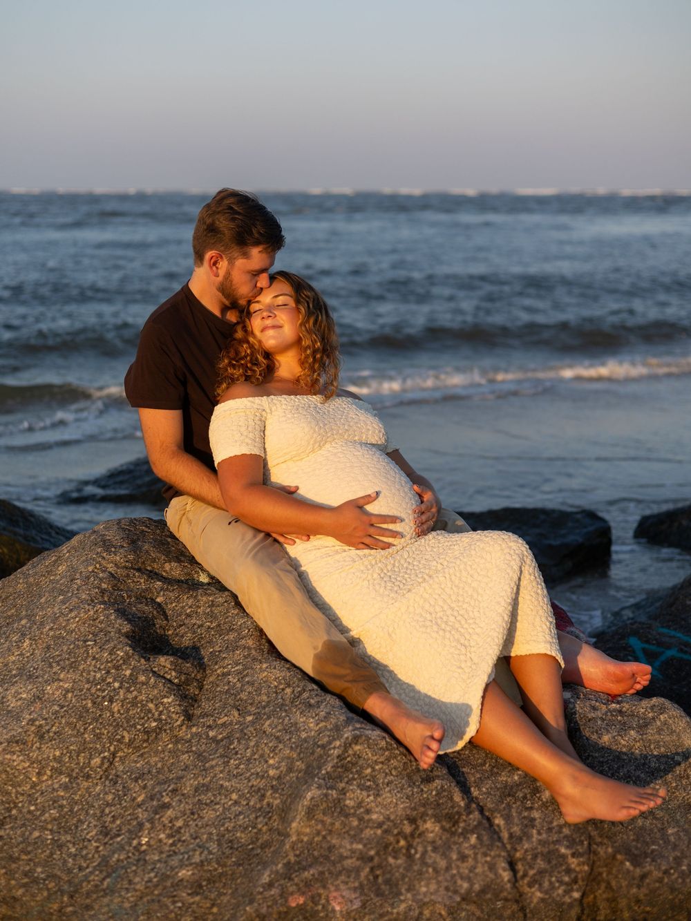 A couple shares a romantic moment together while resting on coastal rocks at sunset by the ocean.
