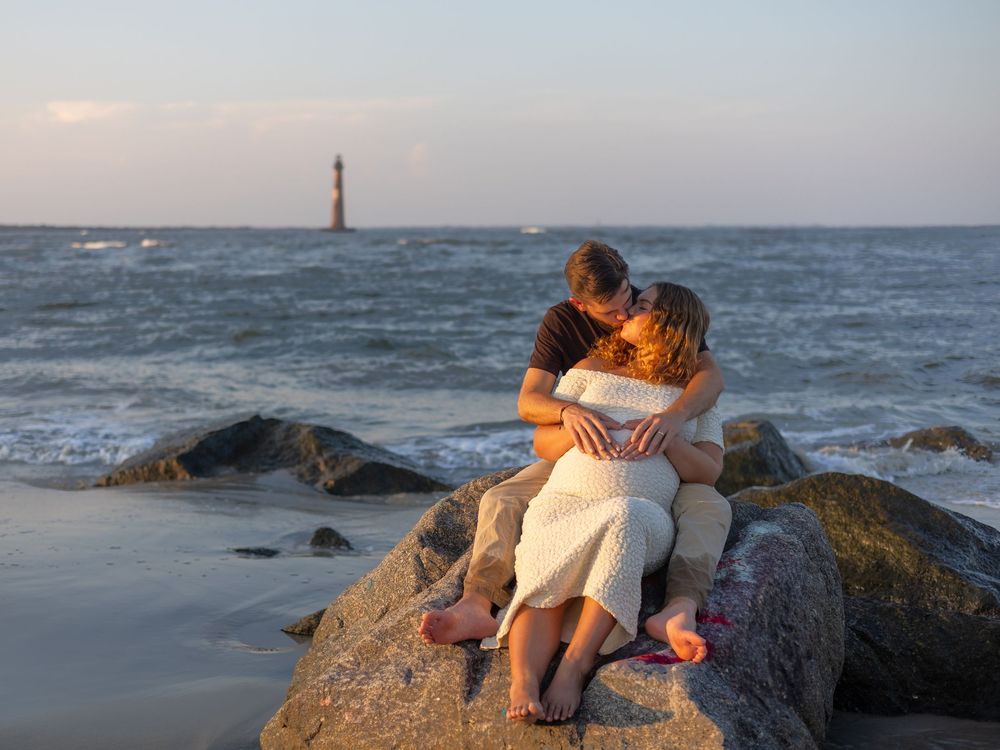 A couple shares a romantic moment on coastal rocks at sunset with ocean waves and a lighthouse in the background.