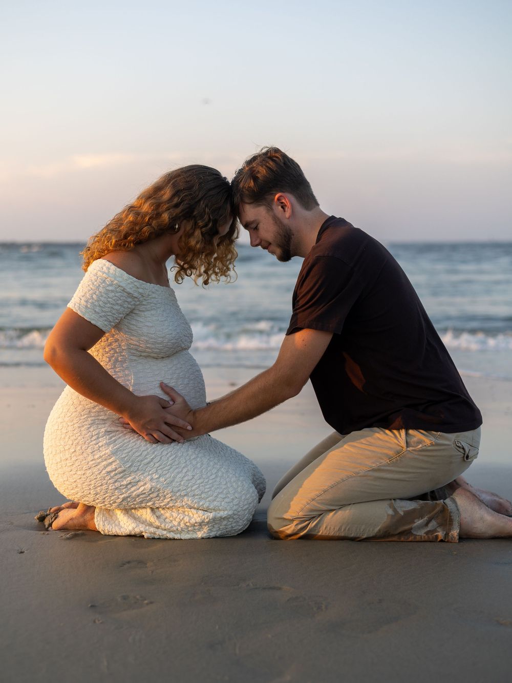 Romantic couple in white clothing sits together on a sandy beach at sunset sharing intimate moments by the ocean.
