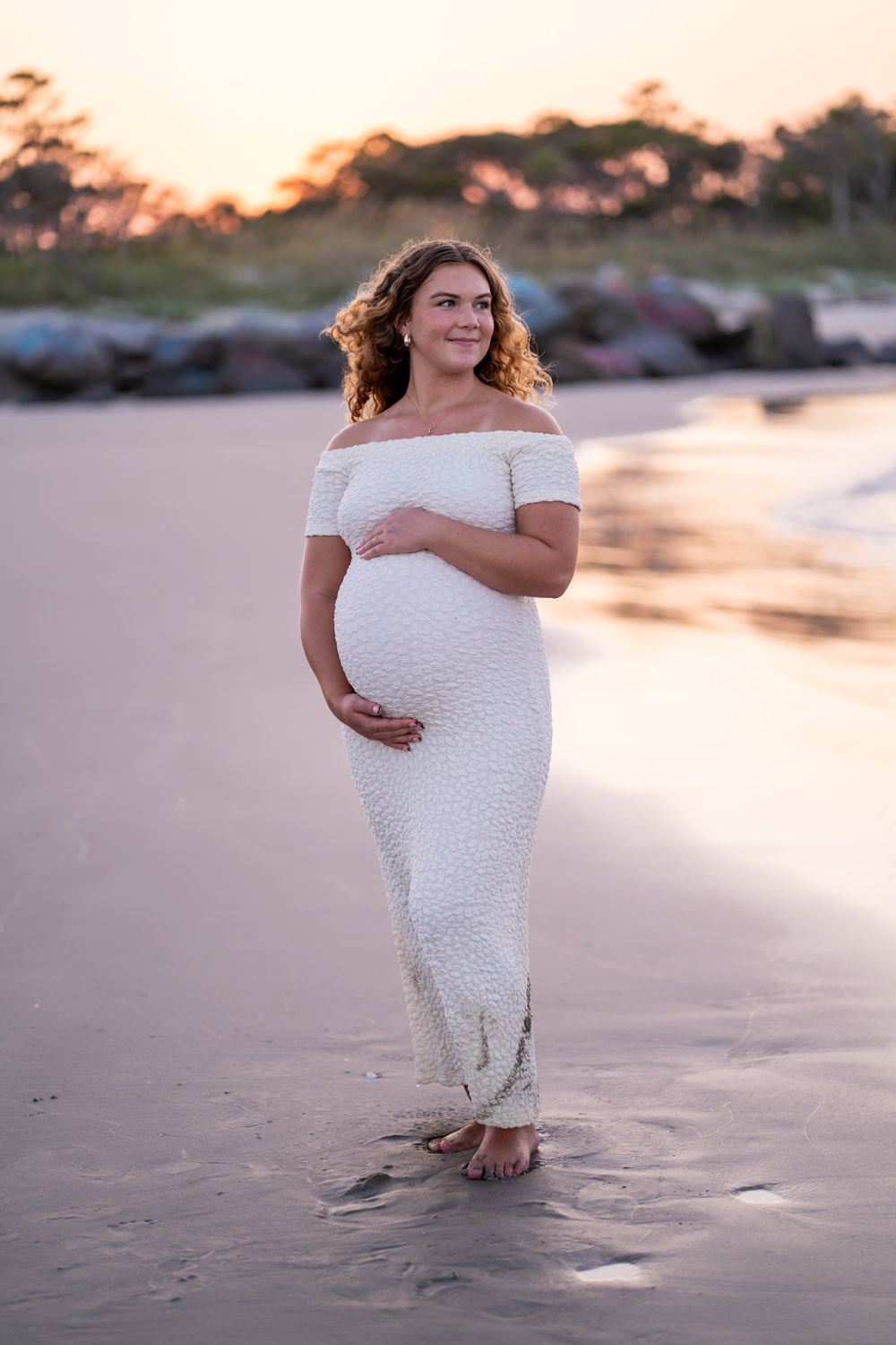 A woman in a white lace dress stands on a beach at sunset with golden light illuminating the scene.