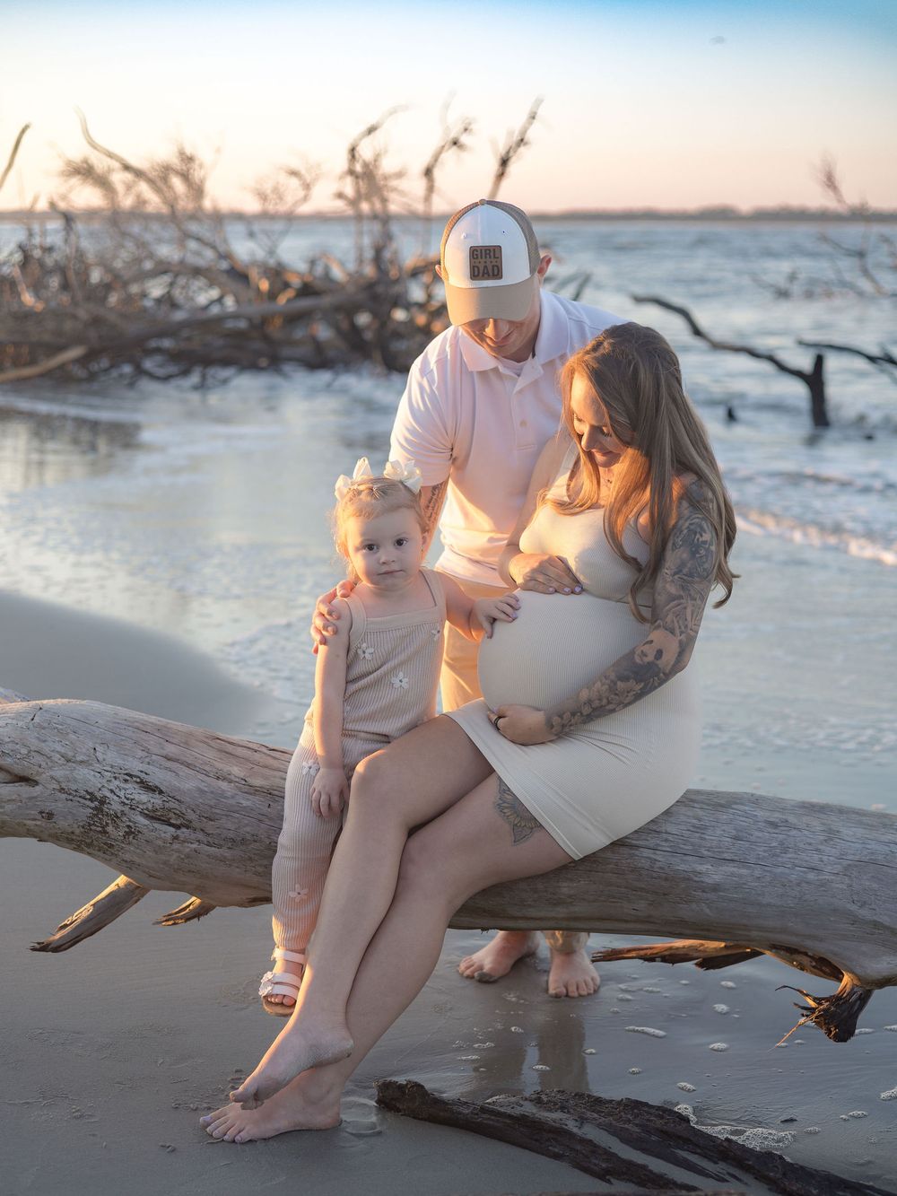 A family sits together on driftwood at the beach during sunset, with gentle waves and coastal vegetation in the background.