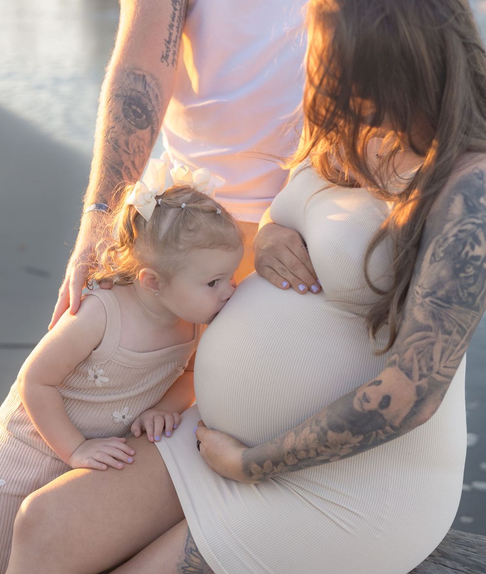 A tender moment captured in sunlight shows a pregnant person in white with tattoos sitting with a small child.