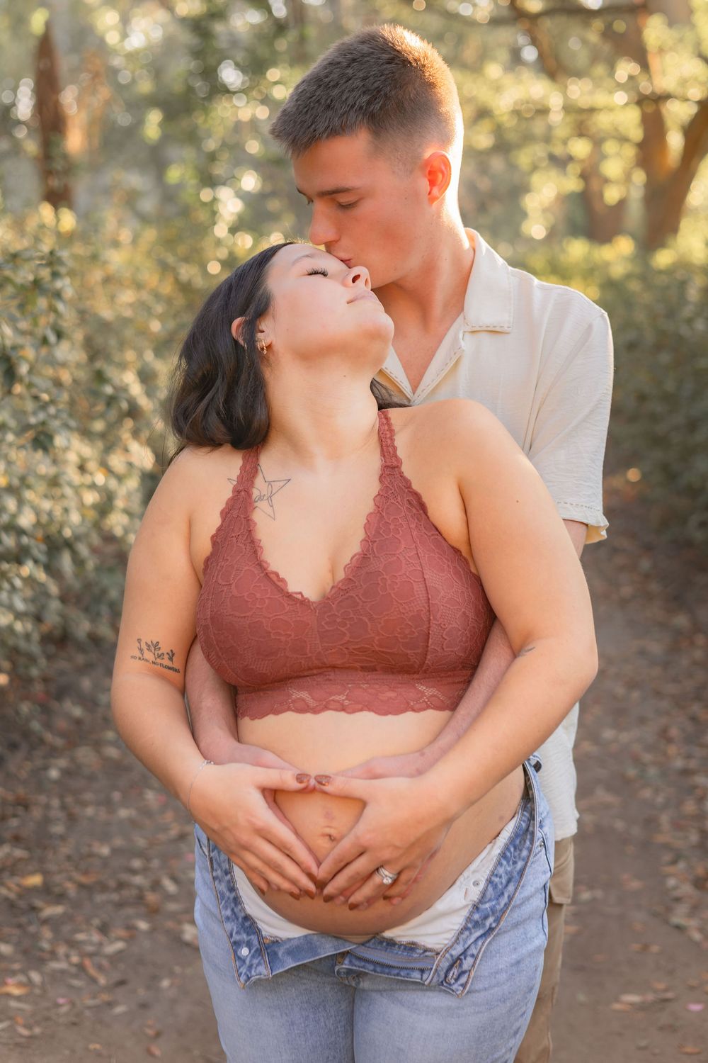 A couple embraces in warm sunset light on a nature path, with golden bokeh through the trees in the background.