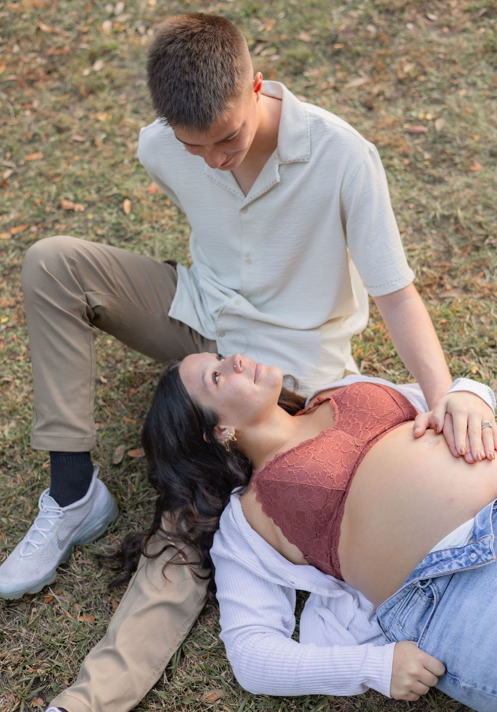 A couple sitting together on grass outdoors, with one person resting their head on the other's lap in casual clothing.