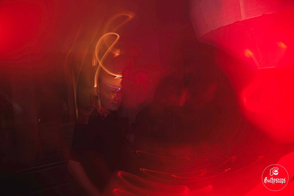 Atmospheric red lighting illuminates a silhouetted figure performing on stage through a hazy, moody backdrop.