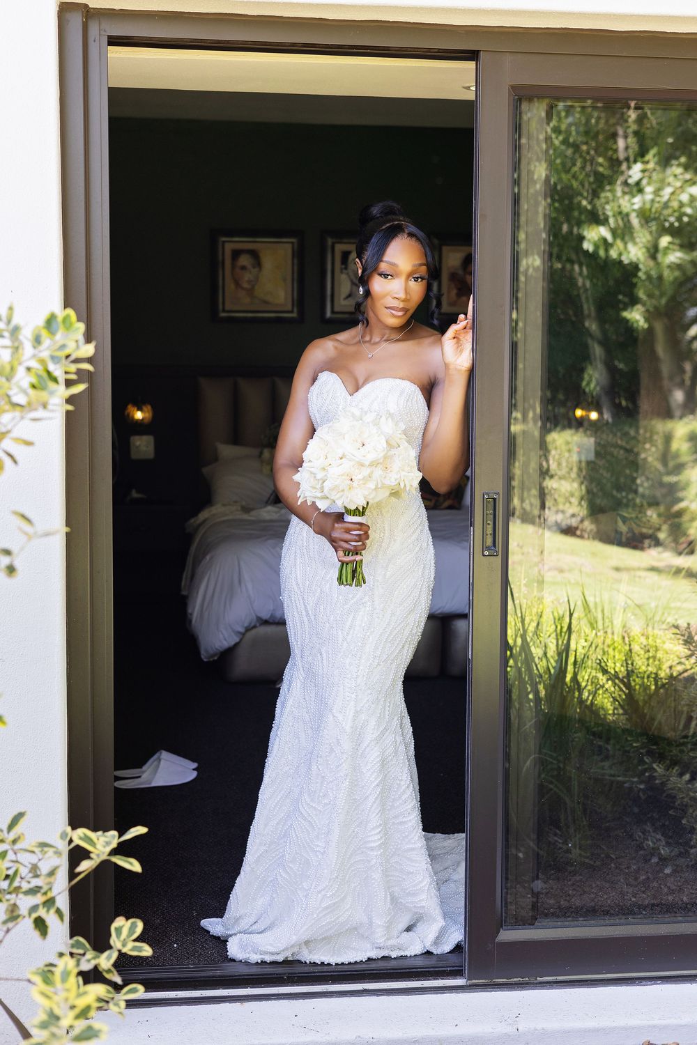 Stunning bride in white lace mermaid wedding gown holds white bouquet while standing in doorway.