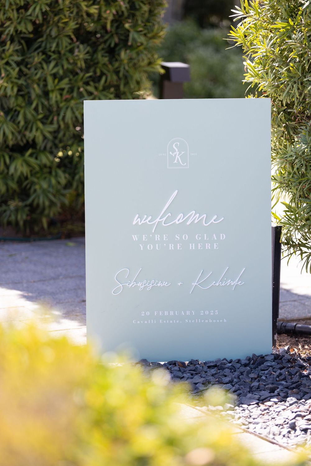 Elegant wedding welcome sign with white text on light blue background displayed in garden setting.