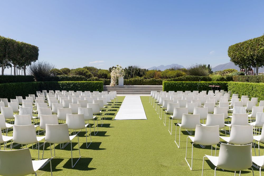 Outdoor wedding ceremony setup with rows of white chairs on green grass and trees in the background.